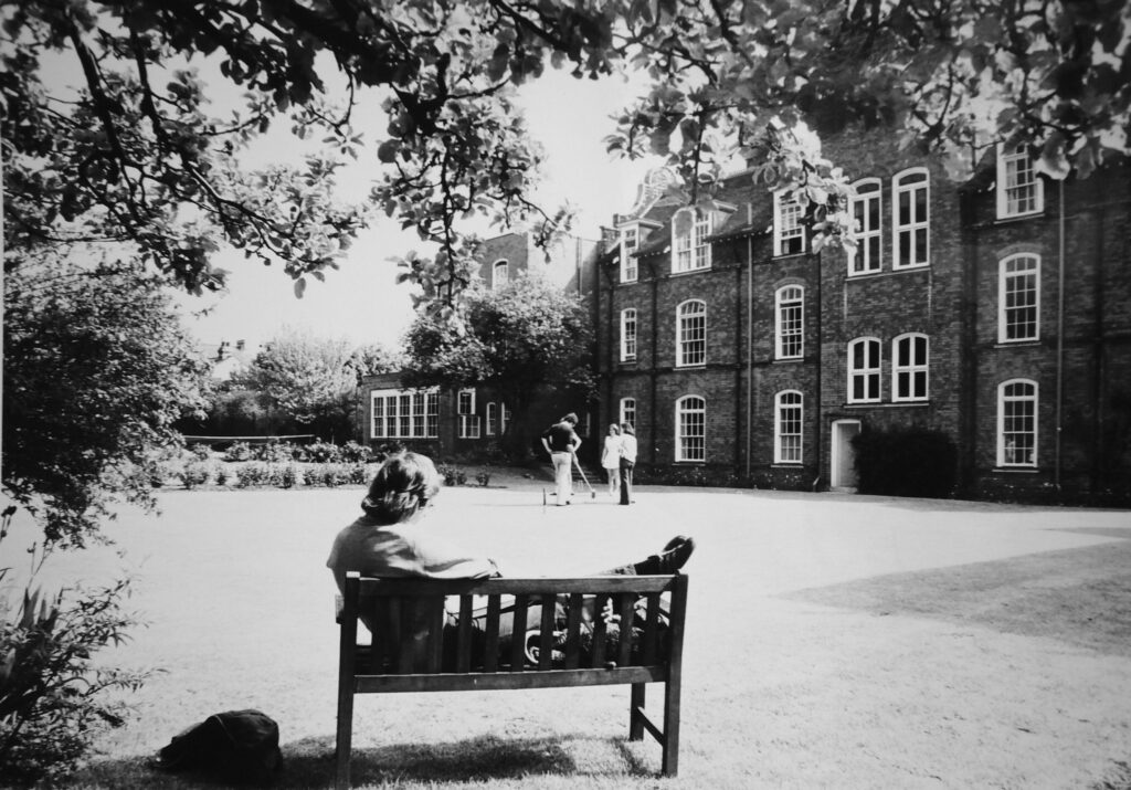 A black and white image of students playing croquet on the back lawn, watched by a student on a bench